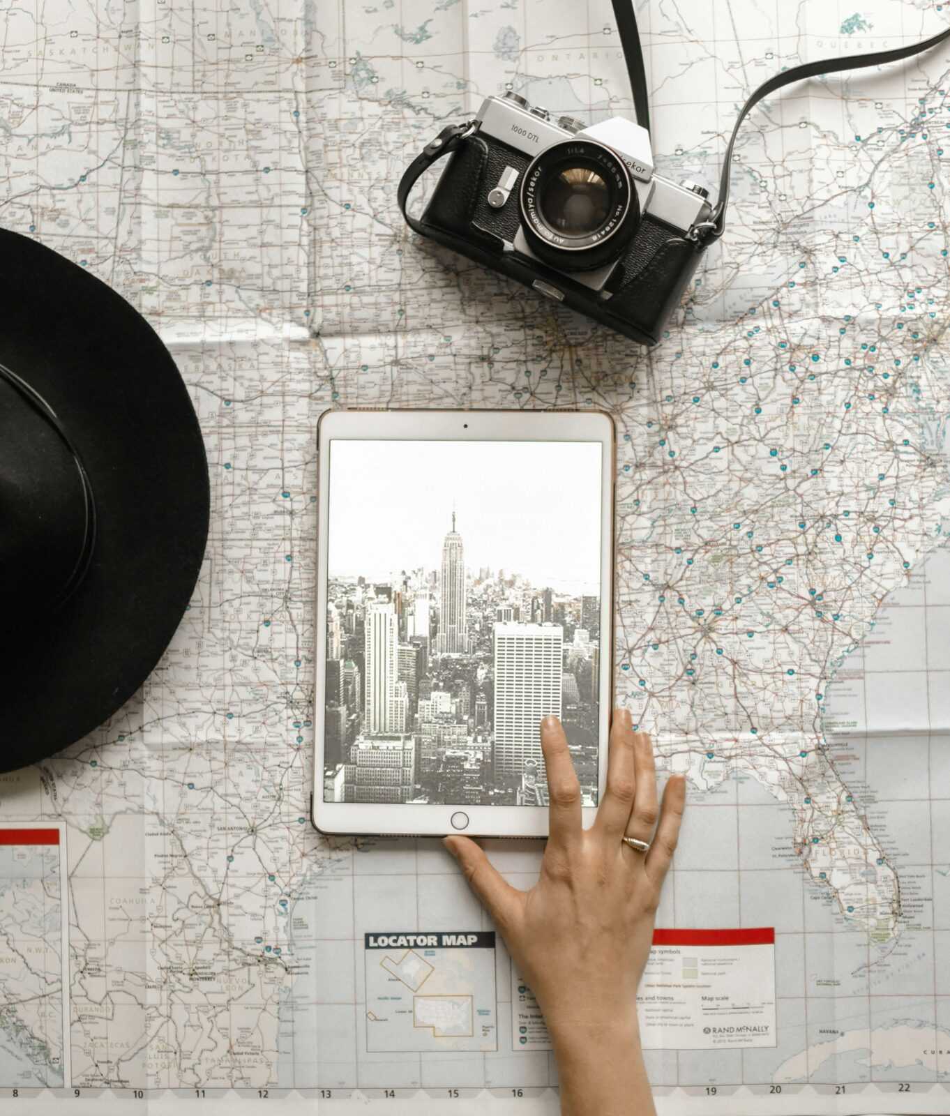 Flat lay of vintage camera, iPad with New York City skyline, and a hand on a map.