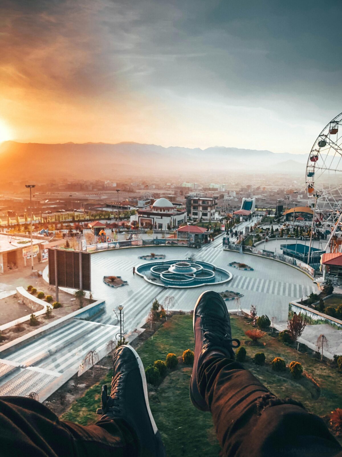 Captivating aerial view of a ferris wheel and urban landscape at sunset with a pair of legs visible.