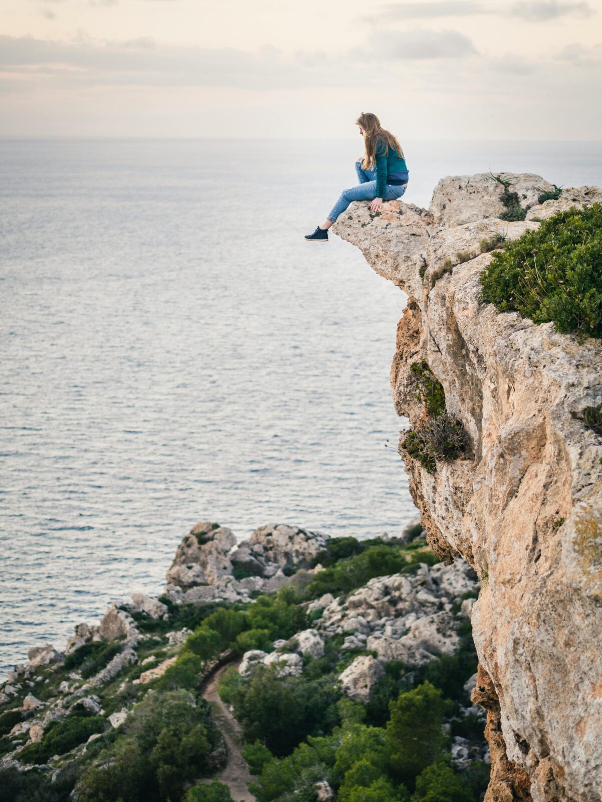 A woman sits daringly on a cliff edge in Mgarr, Malta, overlooking the scenic ocean view.
